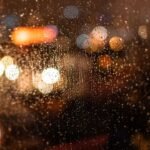 Gotas de chuva no vidro de uma janela à noite, com luzes desfocadas ao fundo