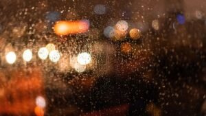 Gotas de chuva no vidro de uma janela à noite, com luzes desfocadas ao fundo