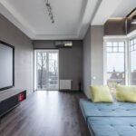 Sala de estar ampla com tela grande na parede e sofá azul junto às janelas