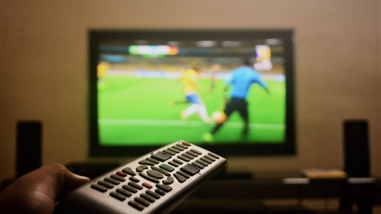 Mão segurando controle remoto diante de uma TV que mostra uma partida de futebol