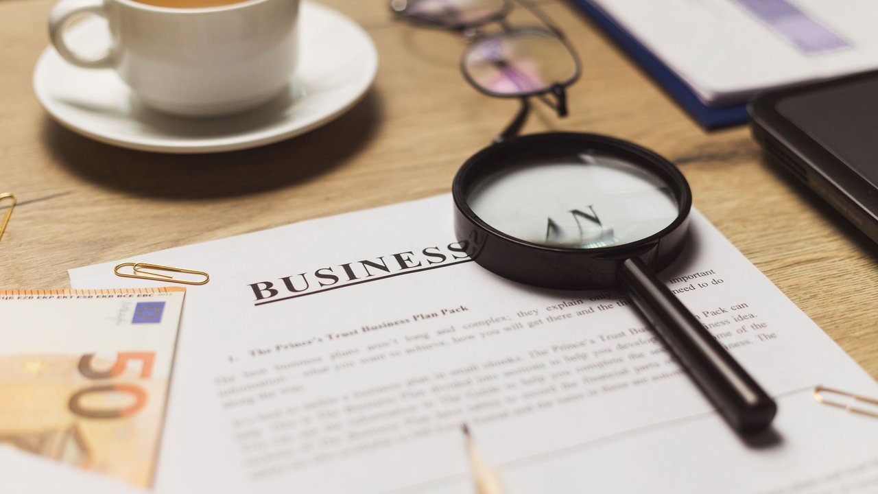 Mesa de trabalho com lupa sobre documentos, xícara de café e cédulas