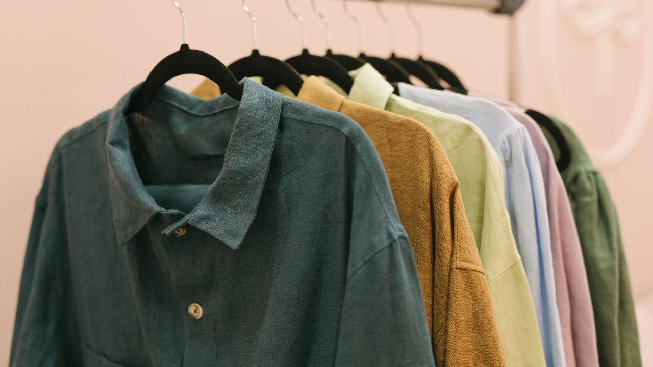 Camisas e casacos em tons de verde, mostarda e rosa pendurados em cabides numa arara