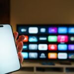 Mão segurando celular diante de uma smart TV com aplicativos na tela