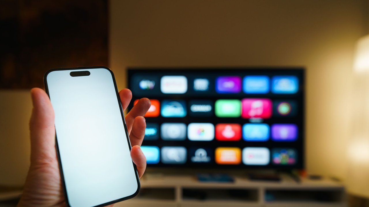 Mão segurando celular diante de uma smart TV com aplicativos na tela